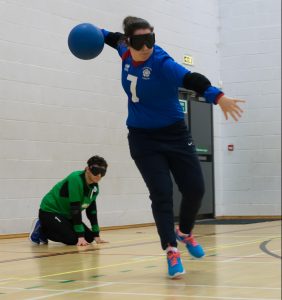 Emma throwing a goalball during an event. Emma's wearing her South Yorkshire shirt mid throw, her right arm is pulled back ready to push through.