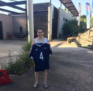 Emma standing on a path with her Great Britain navy blue shirt with the number 4 on it. Emma is smiling as she is holding her shirt up.