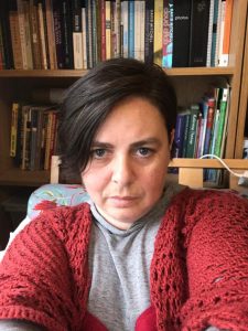Frin Lewis-Smith taking a selfie. Frin has short black hair and is wearing a red cardigan with a grey t-shirt. There is a bookcase behind her.