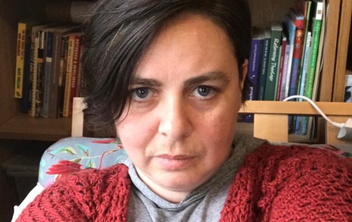 Frin Lewis-Smith taking a selfie. Frin has short black hair and is wearing a red cardigan with a grey t-shirt. There is a bookcase behind her.