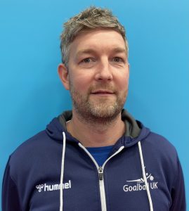 Gary Fraser having a photo taken showing his face whilst wearing a navy blue Goalball UK jacket as he is standing in front of a turqouise background.
