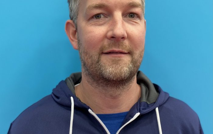 Gary Fraser having a photo taken showing his face whilst wearing a navy blue Goalball UK jacket as he is standing in front of a turqouise background.