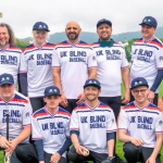 UK Blind Baseball team photo at a tournament in Italy. British Blind Sport staff member Alex Pitts is with the team who are all wearing their prodominately white jerseys with blue writing.