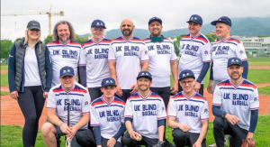 UK Blind Baseball team photo at a tournament in Italy. British Blind Sport staff member Alex Pitts is with the team who are all wearing their prodominately white jerseys with blue writing.