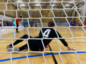 Stuart Hudson playing for RNC Academy on court wearing the number 4 jersey. The image is taken from behind Stuart as he s in the ready positon, with the camera peeking through the goal netting.