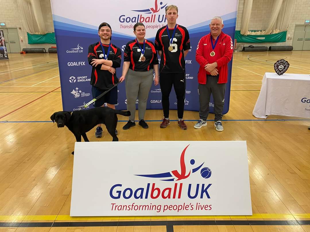 York St John novice shield team standing together in front of a Goalball UK pop up banner. 3 players are wearing their black jerseys with coach Rob Avery wearing his referee jacket. Evie the guide dog has joined the team photo.