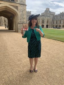 Emma Evans standing on a path in a black and green dress holding her MBE in her right hand, and pointing towards it with her left hand with a big smile!