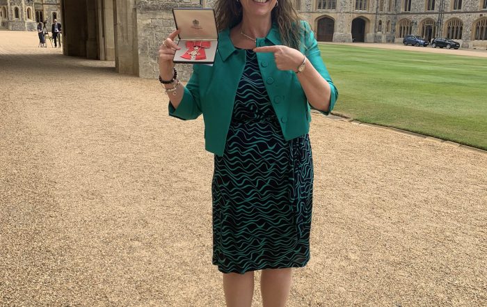 Emma Evans standing on a path in a black and green dress holding her MBE in her right hand, and pointing towards it with her left hand with a big smile!