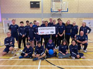 #FindTheNext Goalball Academy team photo in a sports hall. 21 people are joining in for the photo, all wearing navy blue Goalball Academy shirts. Alex Ulysses is holding a sign in the middle which reads "I'm proud to be part of the #FindTheNext Goalball Academy!"