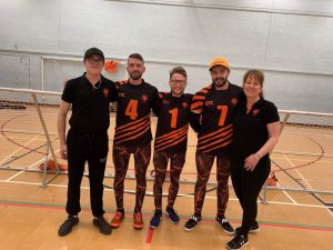 Fen Tigers group photo in front of a goalball goal. Left to right: Assistant coach Daniel Beddis, Dom, Joe, Dan Roper, and Head Coach Emma Evans. Everyone is wearing some form of Fen Tigers kit, weather that is playing kit for Dom, Joe, and Dan, or polos for Daniel and Emma.