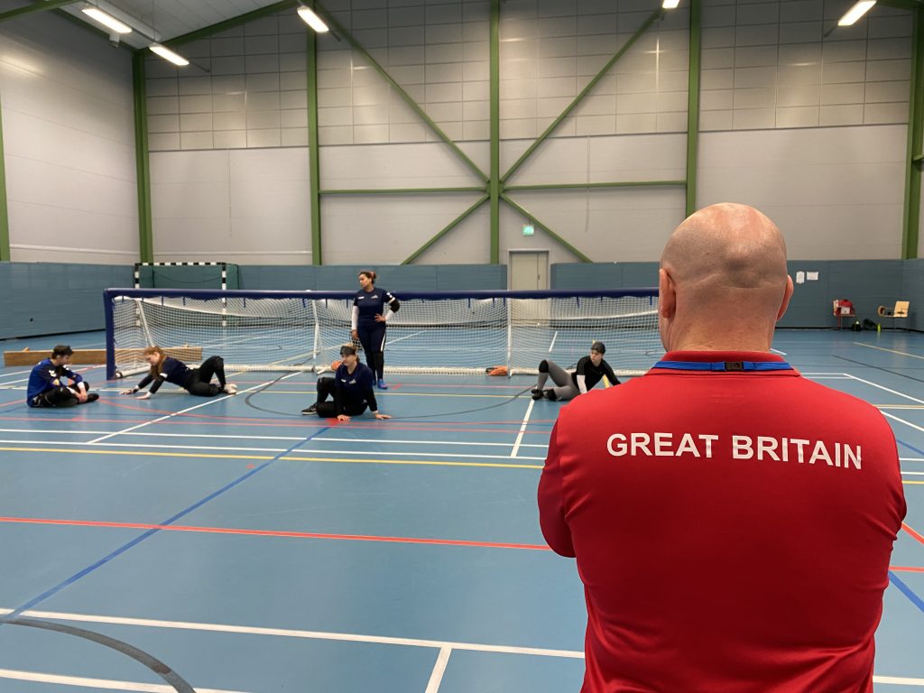 GB Women training at the European Para Youth Games. All players are on one side of the court in positions, with coach Tommy Britain onlooking wearing a red GB top.