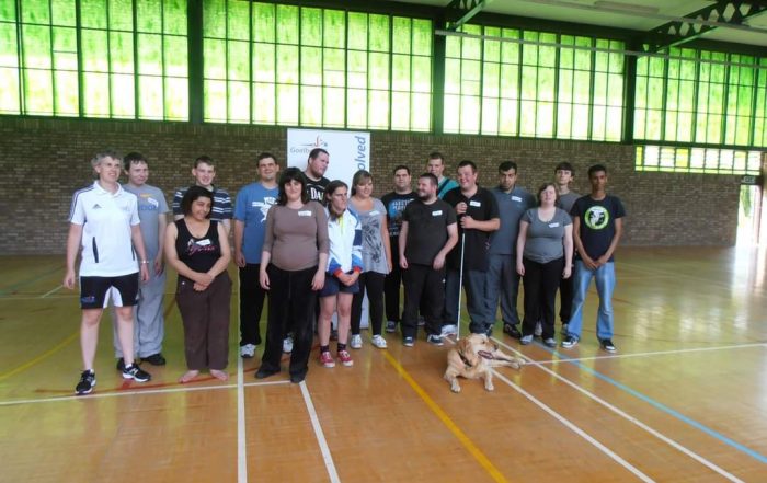A group photo of people attending a past VICTA youth camp, everyone has taken part in a goalball session led by Kathryn Fielding. Behind them is window panels which is giving off a green effect, looking down onto a wooden sports hall floor.