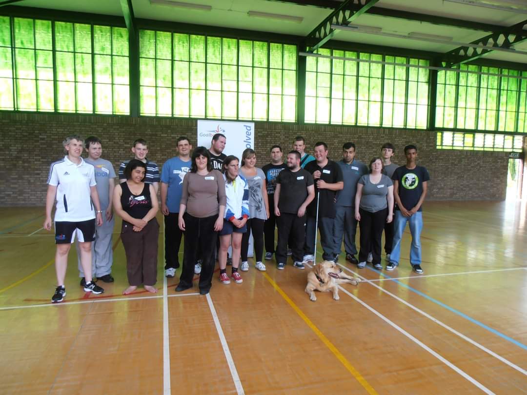 A group photo of people attending a past VICTA youth camp, everyone has taken part in a goalball session led by Kathryn Fielding. Behind them is window panels which is giving off a green effect, looking down onto a wooden sports hall floor.