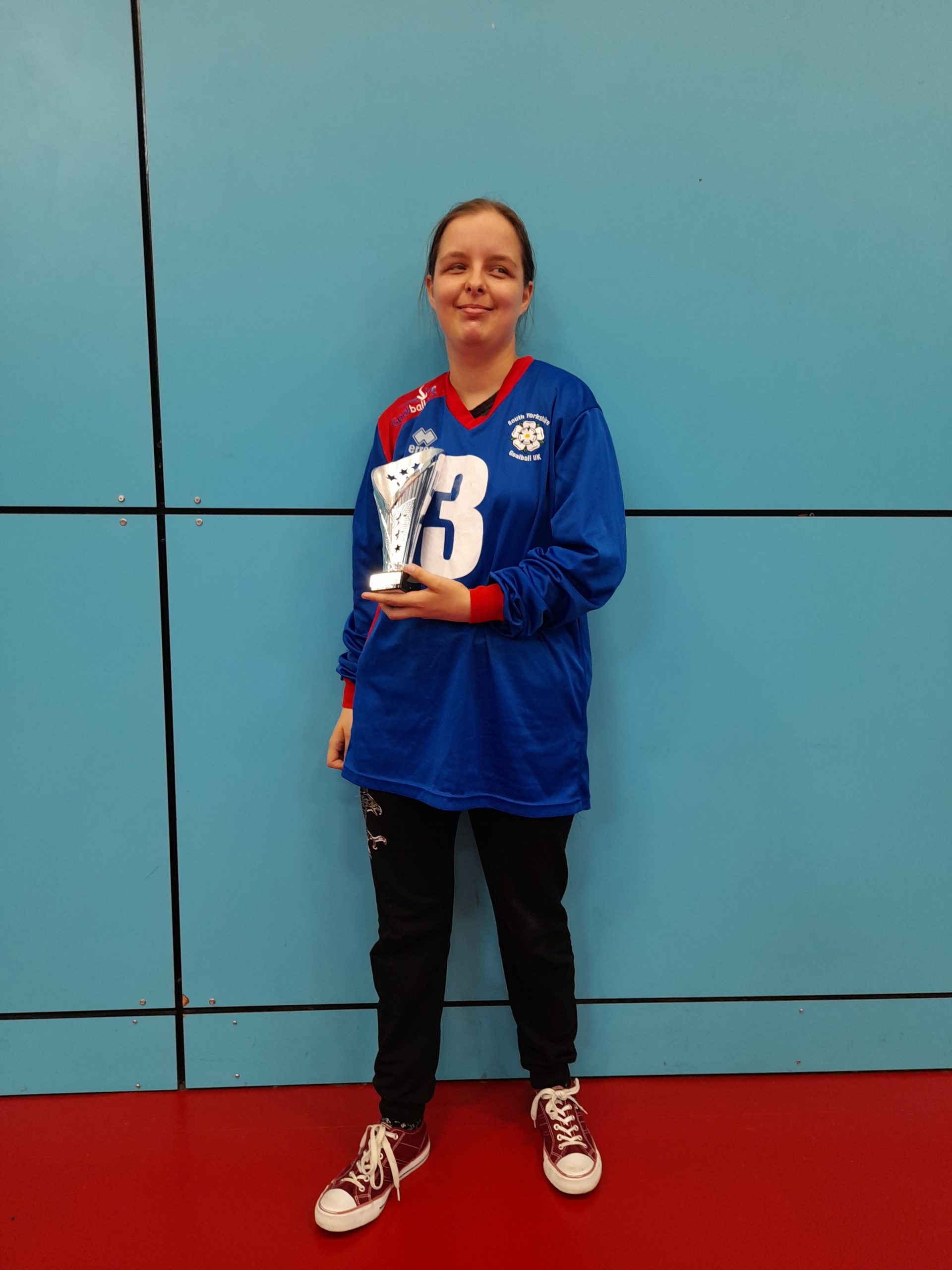 Klaudia Orgel standing in a blue South Yorkshire goalball jersey, and black tracksuit bottoms holding a silver trophy.