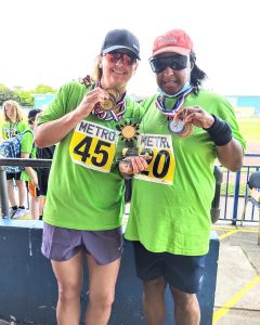 Susan standing with a friend, who are both wearing bright green athletics tops and holding up their medals they have won at a Metro Blind Sport athletics event.