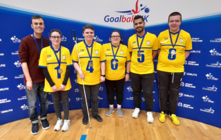 Group photo of six West Yorkshire goalball team members. They are all stood infront of a GoalballUK banner. 5 of the team are wearing yellow team tops and one is wearing a jumper