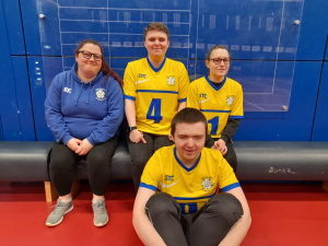 A group of three people sat on a bench with one person sat on the floor infront. three of the people are wearing yellow west yorkshire goalball team tops and one is wearing a blue hoodie.