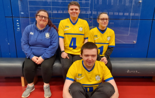 A group of three people sat on a bench with one person sat on the floor infront. three of the people are wearing yellow west yorkshire goalball team tops and one is wearing a blue hoodie.
