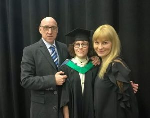 Ella on graduation stood with two older adults. Ella is wearing a graduation gown and cap and the man is wearing a suit, the woman is wearing a black dress.