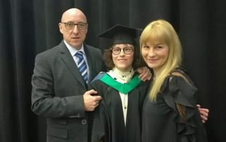 Ella on graduation stood with two older adults. Ella is wearing a graduation gown and cap and the man is wearing a suit, the woman is wearing a black dress.