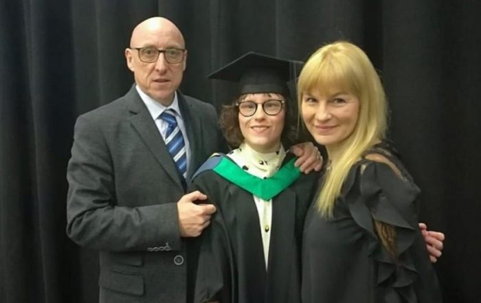 Ella on graduation stood with two older adults. Ella is wearing a graduation gown and cap and the man is wearing a suit, the woman is wearing a black dress.