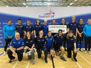 A #FindTheNext Goalball Academy team photo, with everyone wearing navy blue or light blue shirts and jumpers. The team is in two rows, the first row kneeling, with the top row standing.