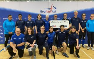 A #FindTheNext Goalball Academy team photo, with everyone wearing navy blue or light blue shirts and jumpers. The team is in two rows, the first row kneeling, with the top row standing.