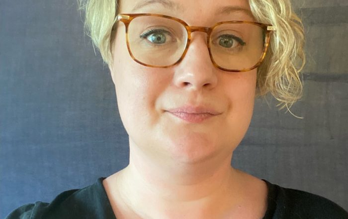 A head and shoulders shot of Jo. She is wearing a black v neck jumper. She has short, blonde hair and is wearing glasses.