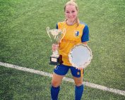 Kirsty Allen having her photo taken on an artificial football pitch in her yellow and blue Mansfield kit. Kirsty is holding a silver trophy in her right hand, and a silver trophy plate in her left hand!