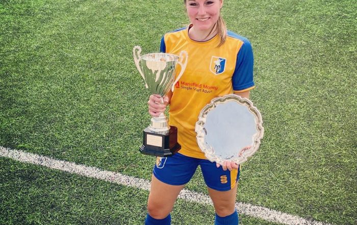 Kirsty Allen having her photo taken on an artificial football pitch in her yellow and blue Mansfield kit. Kirsty is holding a silver trophy in her right hand, and a silver trophy plate in her left hand!