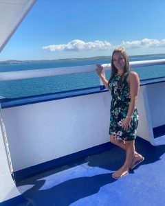 Kirsty Allen standing on deck of a commercial boat in a floral dress. There are bright blue skies behind her, and blue water for miles!