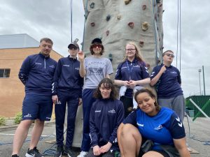 Team Macarana coming together for a team photo in front of a mobile rock climbing wall. 7 people are stood together, smiling together, with 6 of the 7 wearing navy blue Goalball UK shirts!