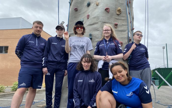 Team Macarana coming together for a team photo in front of a mobile rock climbing wall. 7 people are stood together, smiling together, with 6 of the 7 wearing navy blue Goalball UK shirts!