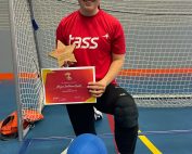 Megan Smithson-Booth wearing a red TASS top and black tracksuit bottoms. Megan is knelt down with her left leg forward, holding her TASS most determined award plaque.