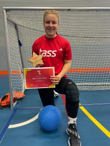 Megan Smithson-Booth wearing a red TASS top and black tracksuit bottoms. Megan is knelt down with her left leg forward, holding her TASS most determined award plaque.