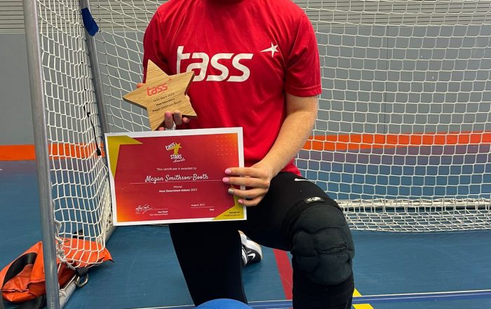 Megan Smithson-Booth wearing a red TASS top and black tracksuit bottoms. Megan is knelt down with her left leg forward, holding her TASS most determined award plaque.