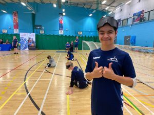 Max wearing his navy blue FindTheNext Goalball Academy Shirt with two thumbs up as a goalball game goes on in the background
