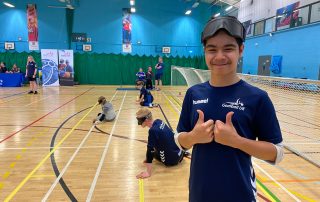 Max wearing his navy blue FindTheNext Goalball Academy Shirt with two thumbs up as a goalball game goes on in the background