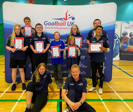 Summer Camp 2022 group photo. Staff members Sophie and Stephen are sat on the sports hall floor, with the 8 attendees standing behind them with their certificates!