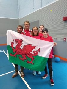 A South Wales group picture in a sports hall at a Hereford competition. two players are holding a big Welsh flag, with 3 people stood closely behind them including Steve Jones.