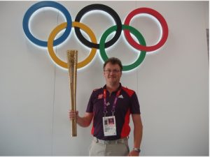 Steve Jones holding the golden Olympic Torch whilst wearing a purple game makers shirt. Steve is standing in front of a wall which has the 5 Olympic Rings.