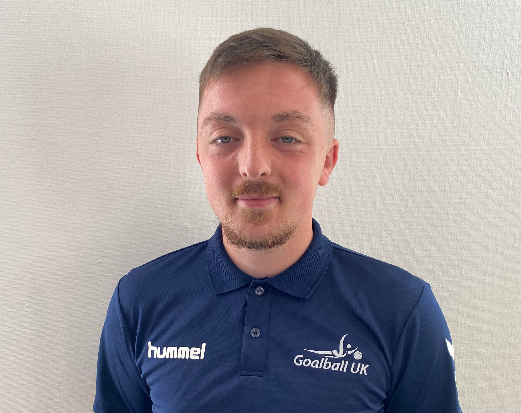 Alex Cockerham portrait. Alex is wearing a blue Goalball UK t-shirt and is stood in front of a white wall