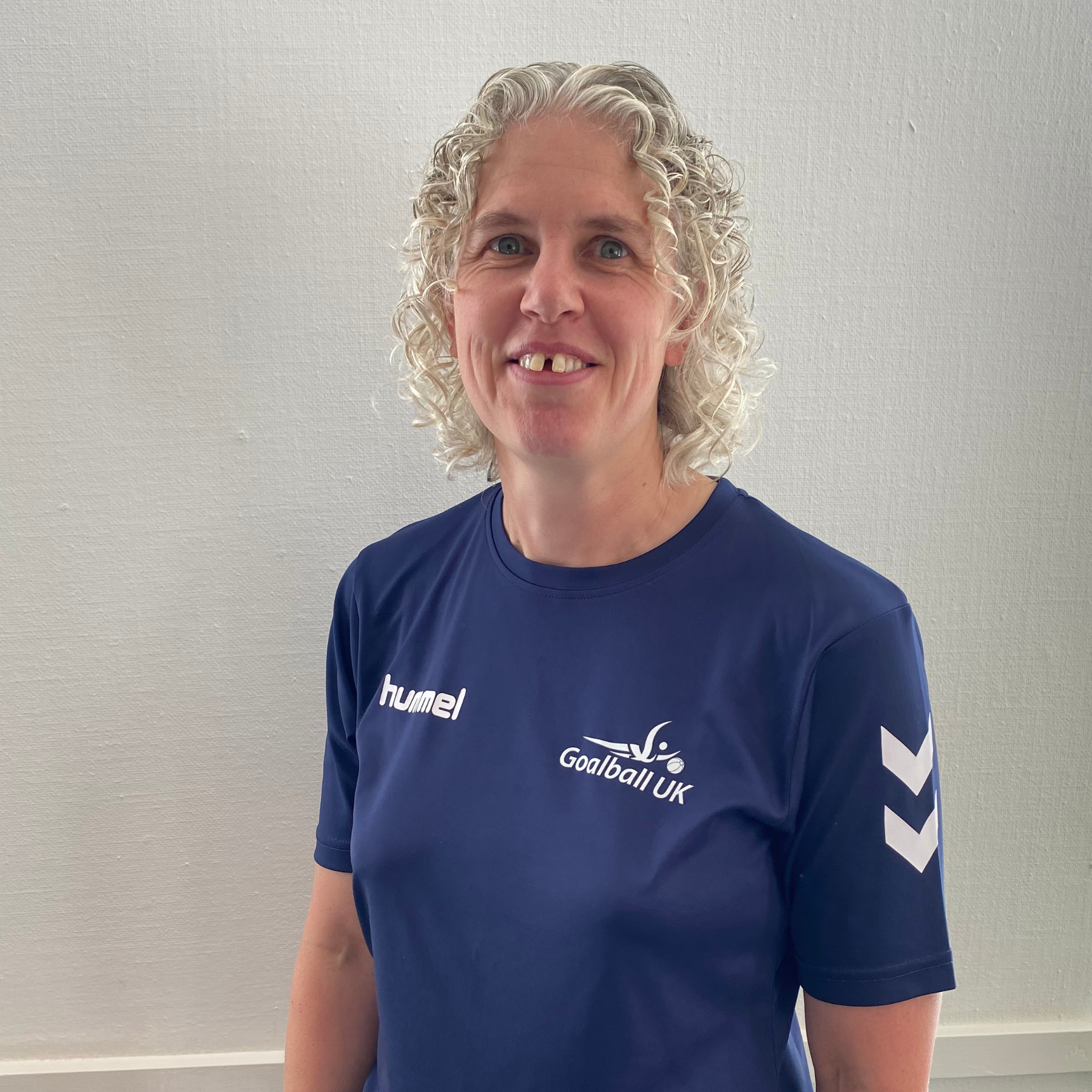 Kathryn Fielding portrait. Kathryn is wearing a blue Goalball UK t-shirt and is stood infront of a white wall