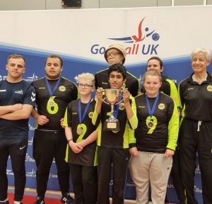 Daniel is stood at the front of a team photo holding a trophy. There are seven other Croysutt Warriors around Daniel. They are stood infront of a Goalball UK banner