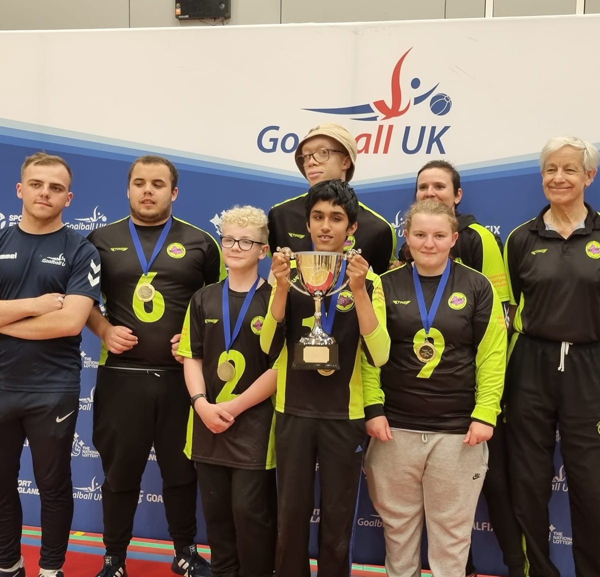 Daniel is stood at the front of a team photo holding a trophy. There are seven other Croysutt Warriors around Daniel. They are stood infront of a Goalball UK banner
