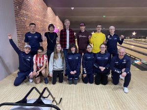 #FindTheNext Goalball Academy team at a bowling alley, with 13 people mixed into two rows in front of a bowling lane. Most of the team are waving their arms in the air!