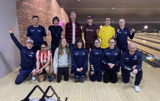 #FindTheNext Goalball Academy team at a bowling alley, with 13 people mixed into two rows in front of a bowling lane. Most of the team are waving their arms in the air!