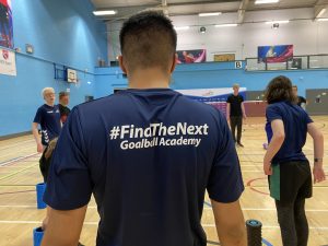 Mustafa Abbas standing in a sports hall during a cool down session with the #FindTheNext Goalball Academy team. He's wearing his navy blue shirt and his back is to the camera, showing white text reading the Academy name.