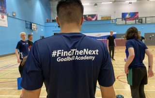 Mustafa taking part in a #FindTheNext Goalball Academy cool down wearing his navy blue shirt. Mustafa is stood up and doing neck shrugs.