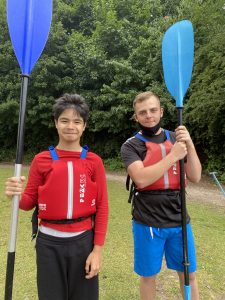 Max and Jack standing together with red life vests on and a paddle each which has a blue end. They're standing on a section of grass with tall green trees in the background, very scenic!